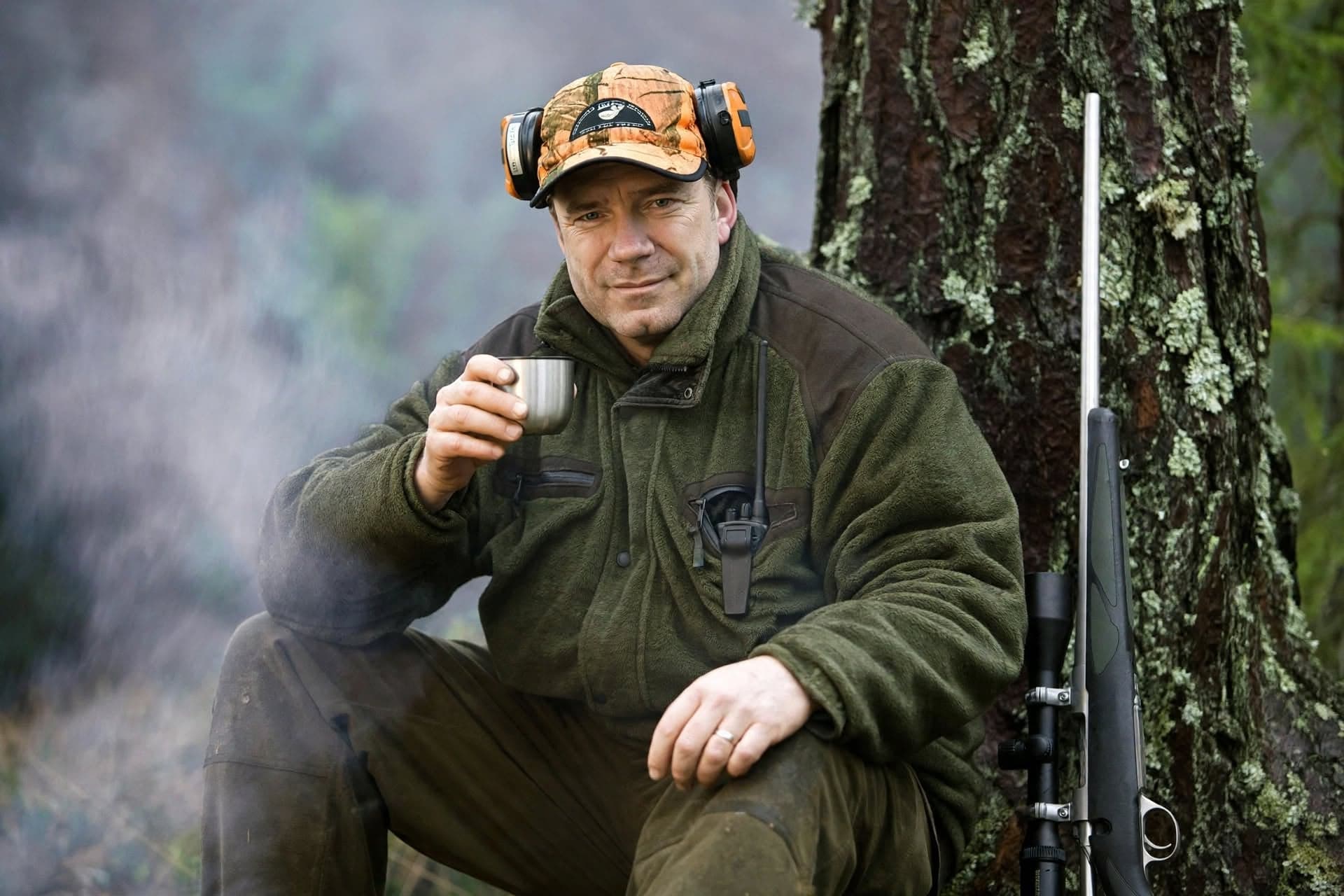 Thomas Ekberg sitter vid en tallstam i skogen med en kaffekopp i handen och kulgeväret lutat mot trädet